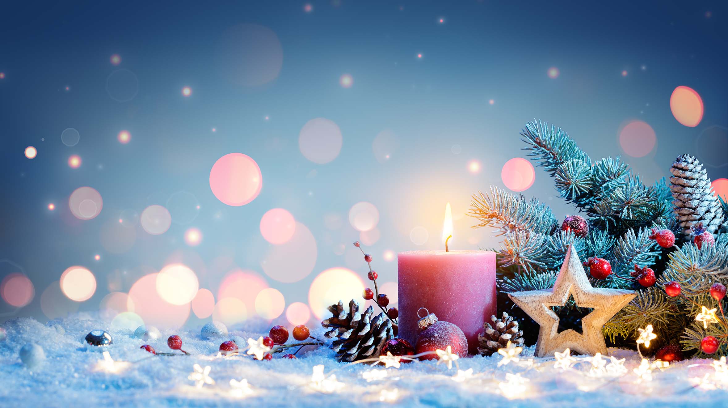 Vor grünen Tannenzeweigen steht eine rote brennende Kerze mit Weihnachtsschmuck.