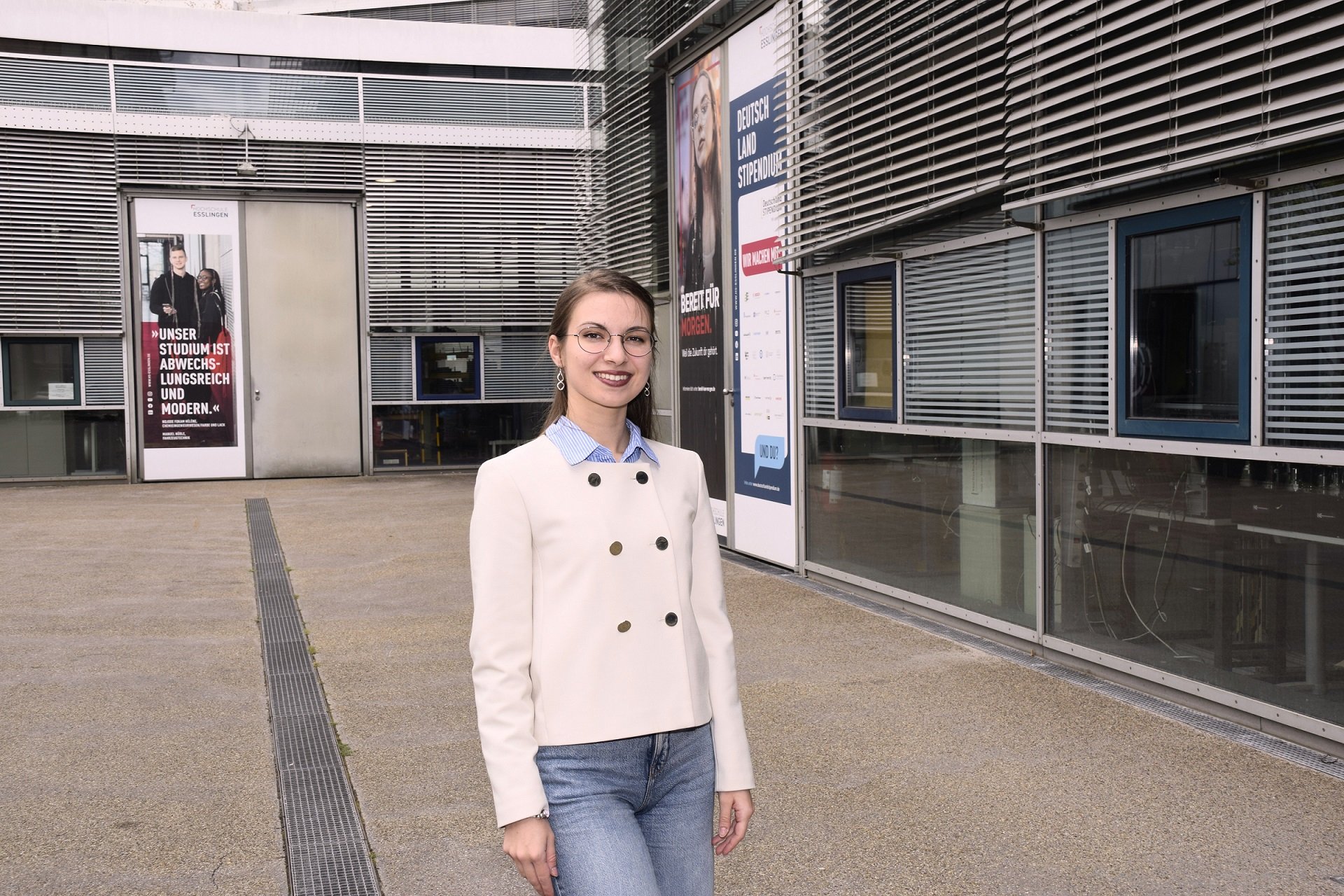 Junge Frau mit Brille und Jacke steht auf dem Campus